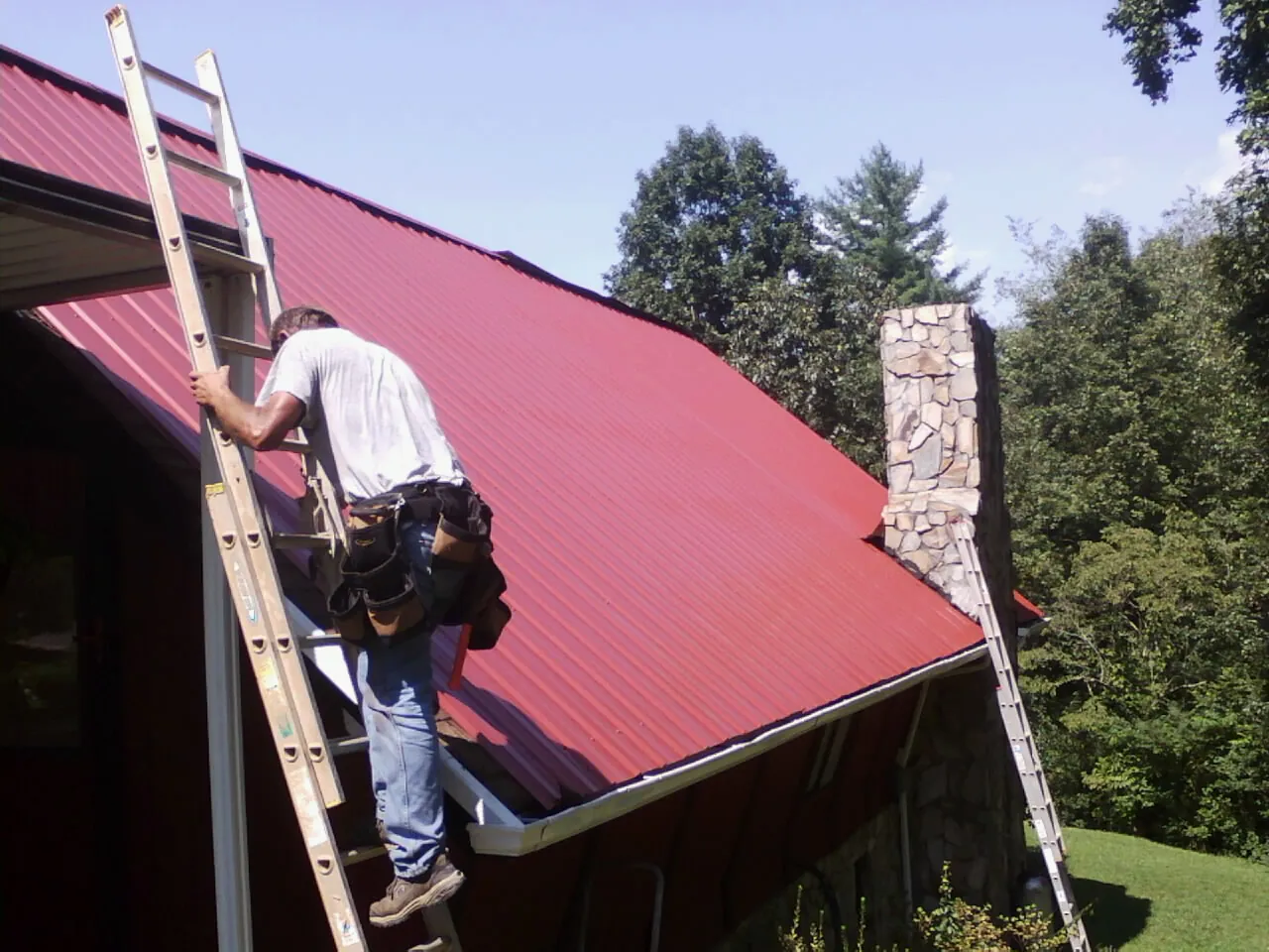Roofer on a ladder beside a red metal roof and stone chimney — Soffit & Fascia Repair by Roofing Brothers in Daisytown